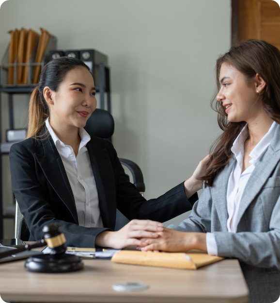 Two professional women sitting at a desk, one comforting the other while holding her hand, with a judge's gavel on the desk.