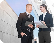 Two business professionals talking while holding documents and a tablet near a modern office building.