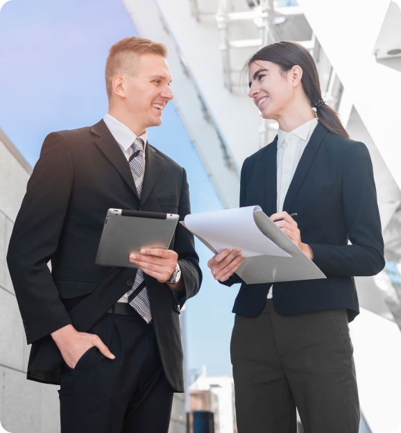 Two business professionals smiling and discussing work outdoors, holding a tablet and clipboard.