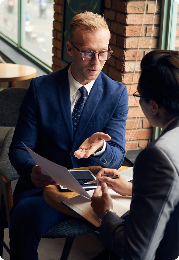 Two businessmen in suits discussing documents at a table in a café with a brick wall background.