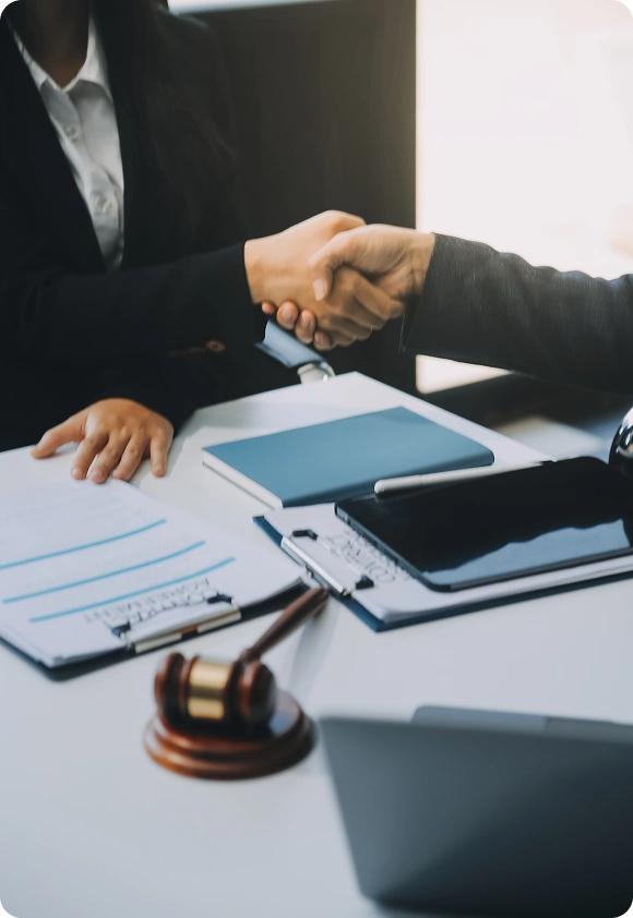 Two professionals in business attire shaking hands over a desk with legal documents, a gavel, and a tablet.