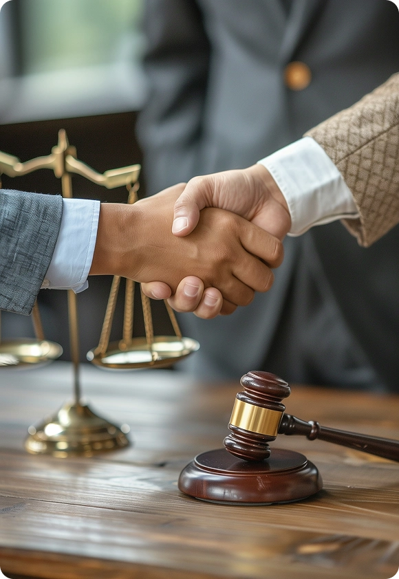 Two people shaking hands in front of a judge's gavel and brass scales of justice on a wooden table.