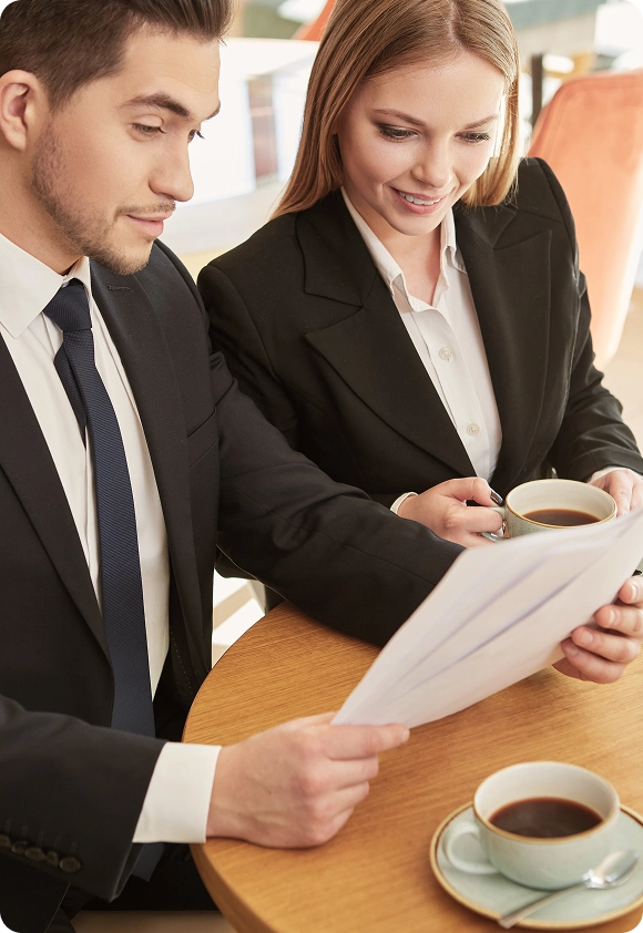 Two business professionals in suits sitting at a table, reviewing documents and drinking coffee.