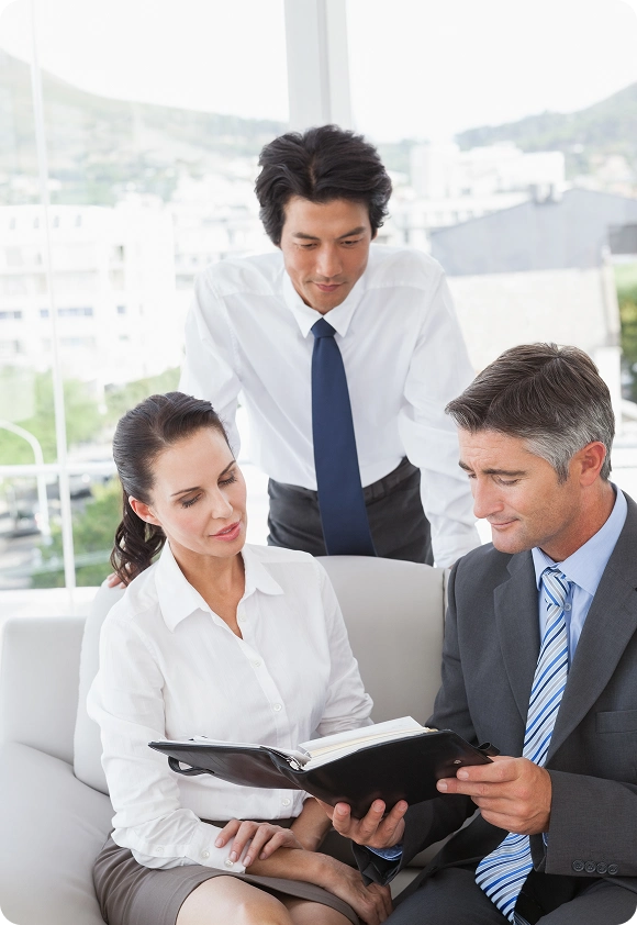 Three business professionals, two men and one woman, reviewing documents together in an office with large windows.
