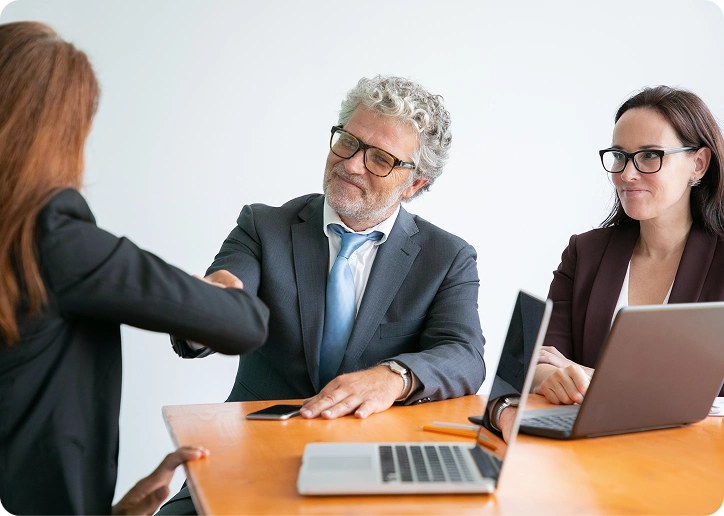 Two businesspeople sitting at a table with laptops, shaking hands with a third person.