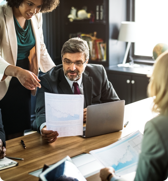 Businessman in suit reviewing a document with charts, while a colleague points at the document, in a modern office meeting.
