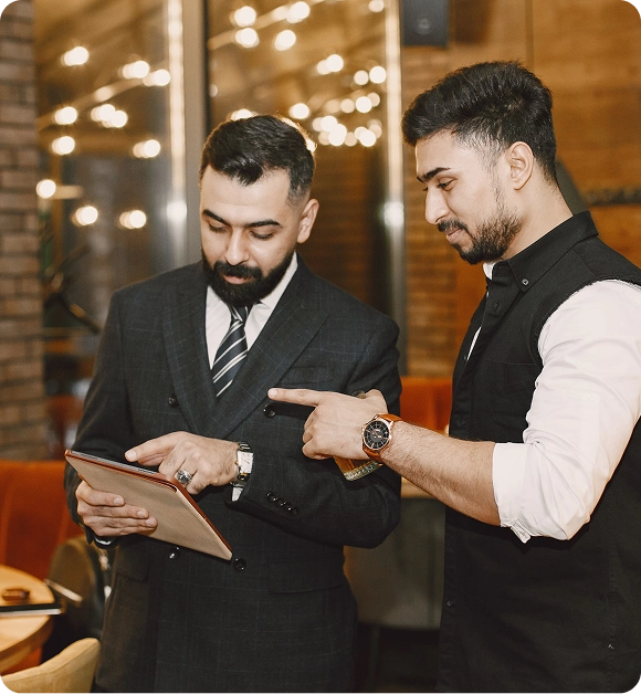 Two men in a warmly lit indoor setting, one wearing a suit and tie holding a tablet, the other pointing at the tablet screen while holding a drink.
