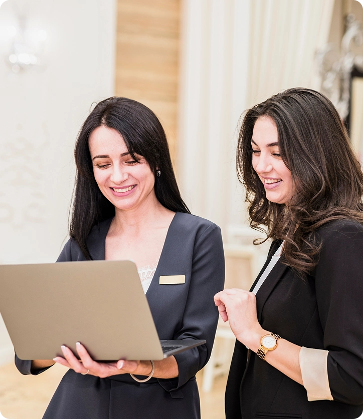 Two women in business attire smiling and looking at a laptop screen together.