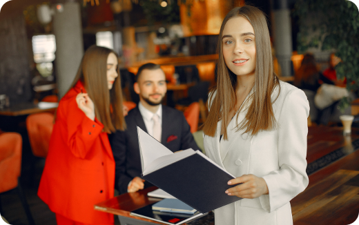 Three young professionals in a modern office; a woman in white holding a folder in the foreground, with a seated man and a woman in red in the background.