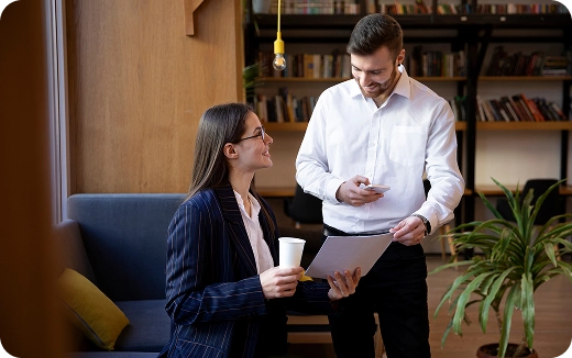 Two colleagues in an office, a woman holding a cup and paper, talking with a standing man holding a phone.