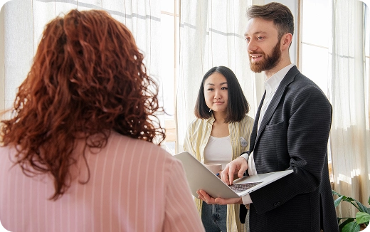 Two people presenting information on a laptop to a woman with curly hair in an indoor setting.