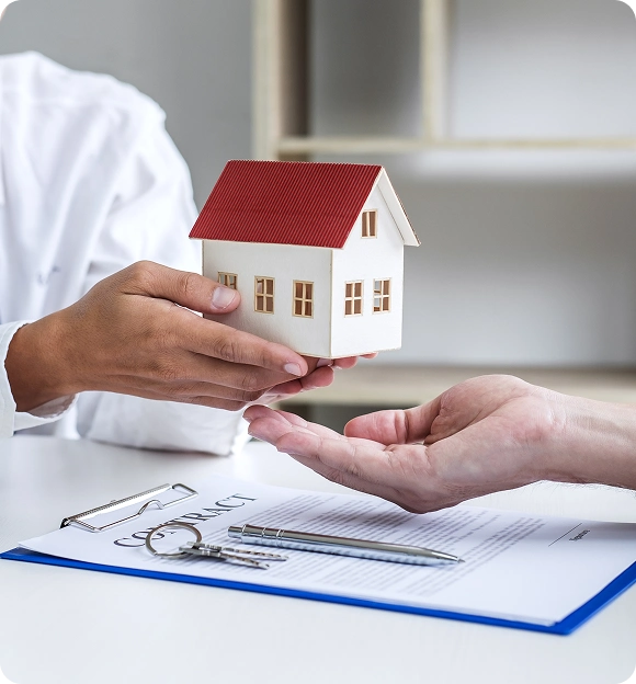 Person handing over a small house model to another person above a contract with keys and a pen on a table.