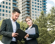 Two business professionals reviewing documents outdoors with high-rise buildings and trees in the background.