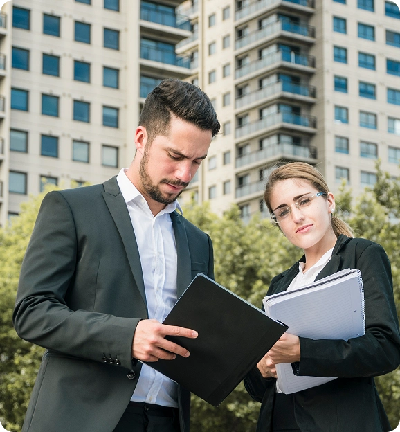 Two business professionals reviewing documents outdoors with office buildings in the background.