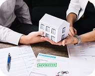 Two people exchanging a small model house over documents, including a paper stamped with 'APPROVED'.