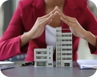 Person in red jacket protecting small model buildings with hands forming a roof shape.