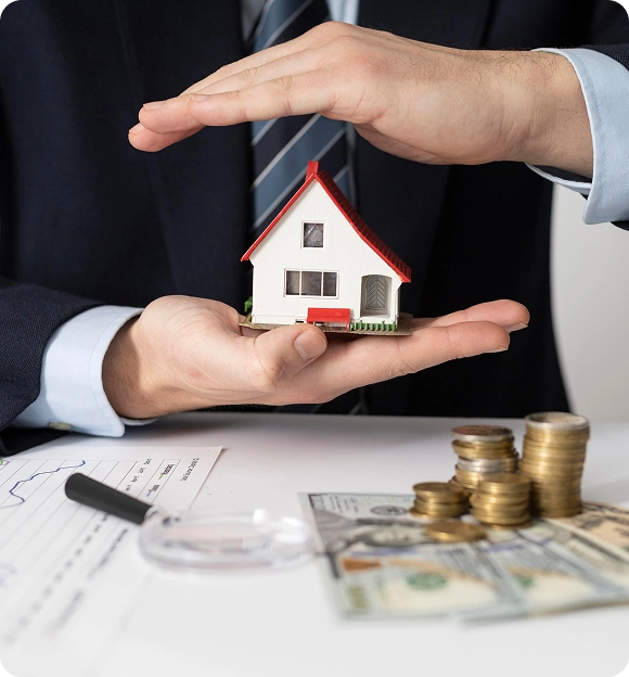Person in suit holding a small model house with one hand underneath and the other hand protecting it, with coins and dollar bills on a table nearby.