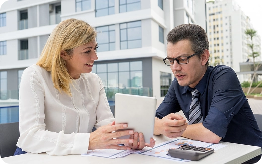 Two business professionals discussing documents and a tablet at an outdoor table with a city building background.