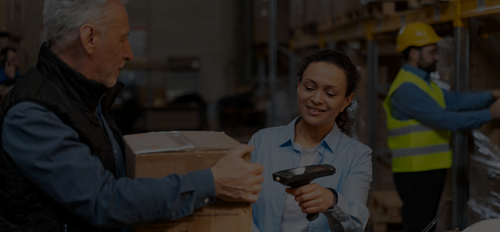 Woman scanning a cardboard box held by a man inside a warehouse with shelves and another worker in a yellow safety vest in the background.
