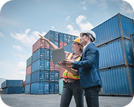 Two workers in safety helmets discussing in front of stacked shipping containers under a blue sky.