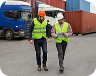 Two men in safety helmets and reflective vests walking and discussing at a shipping container yard.