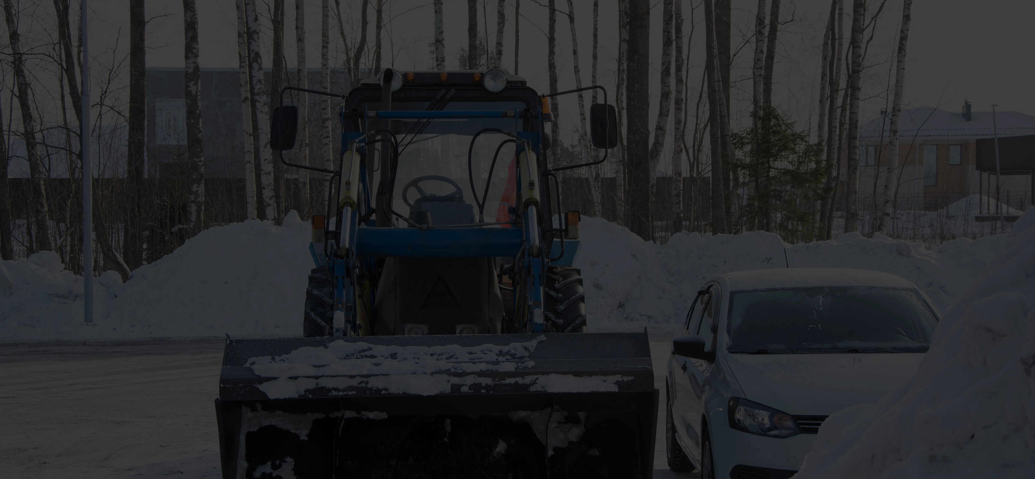 Blue snow plow vehicle with a large blade covered in snow parked next to a white car in a snowy area with trees and houses in the background.