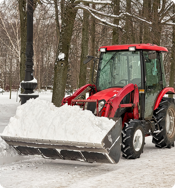 Red snow plow tractor clearing snow on a winter road lined with trees and a lamp post.