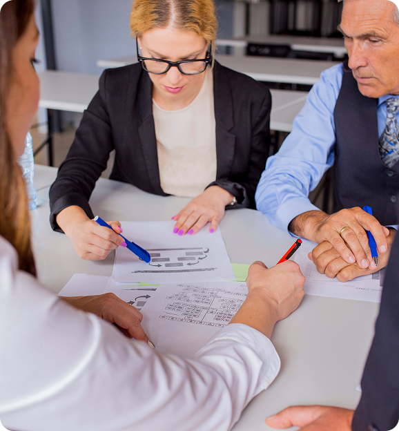 Three people seated around a table reviewing and marking documents and charts with pens during a meeting.