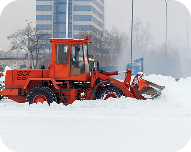 Orange snowplow clearing snow on a street in front of a tall building on a cloudy day.