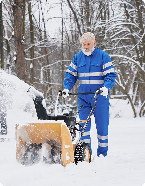 Man in blue reflective workwear operating a snow blower in a snowy wooded area.