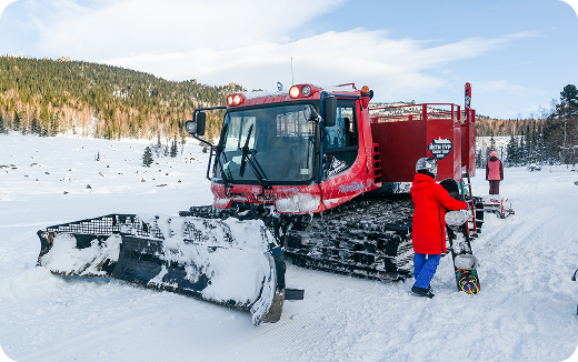 Red snow grooming machine parked on snow with two people in winter gear, one holding a snowboard.