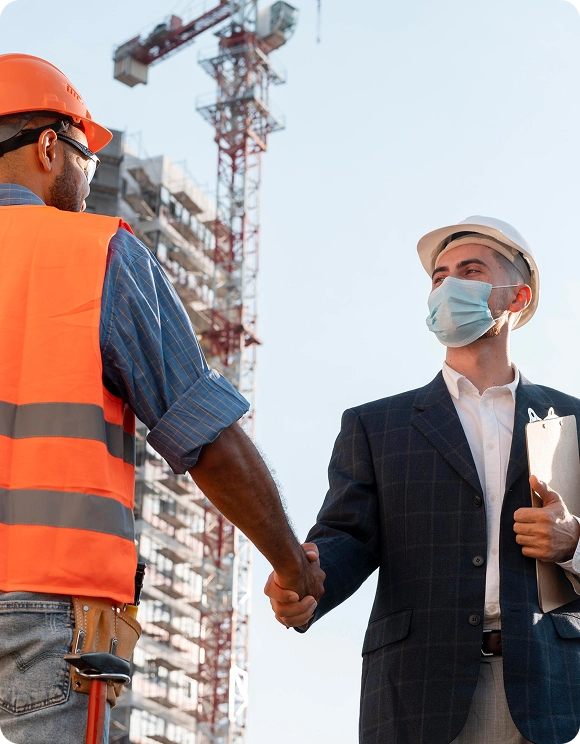 Construction worker in an orange safety vest shaking hands with a businessman wearing a suit, hard hat, and face mask at a building site.