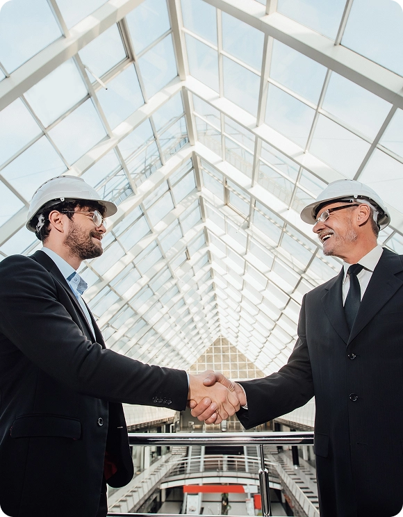 Two men in suits and white construction helmets shaking hands inside a modern glass building atrium.
