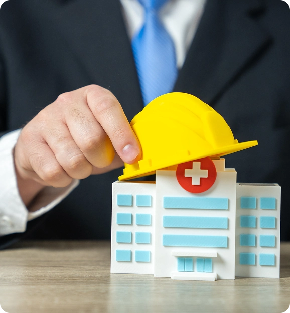 Person in business attire placing a yellow construction helmet on a miniature hospital building model.