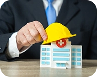 Person in suit placing a yellow safety helmet on a model hospital building.