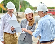 Three construction professionals wearing white safety helmets reviewing plans at a worksite.