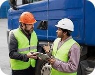 Two construction workers in safety vests and helmets discussing documents beside a blue truck.