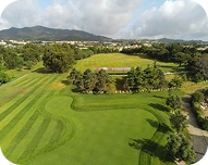 Aerial view of a green golf course with striped fairways, putting greens, trees, and mountains in the background under a cloudy sky.