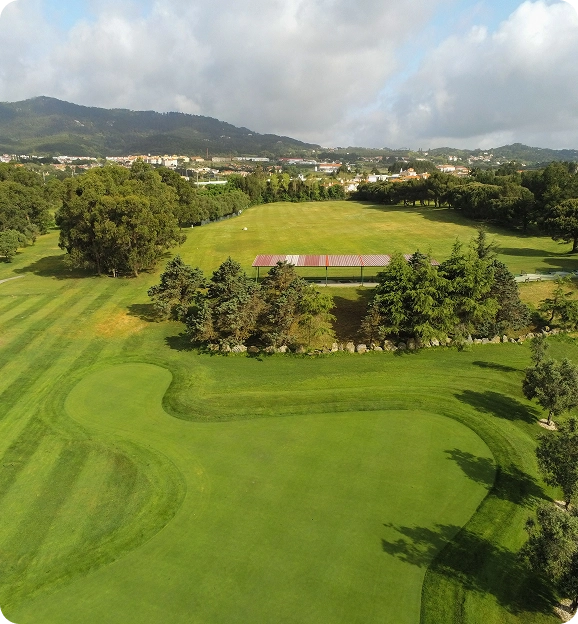 Aerial view of a golf course with a green, sand bunkers, trees, and hills in the background under a partly cloudy sky.
