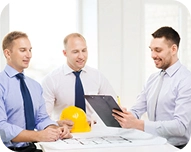 Three men in business attire discussing a blueprint with a yellow construction helmet on the table.