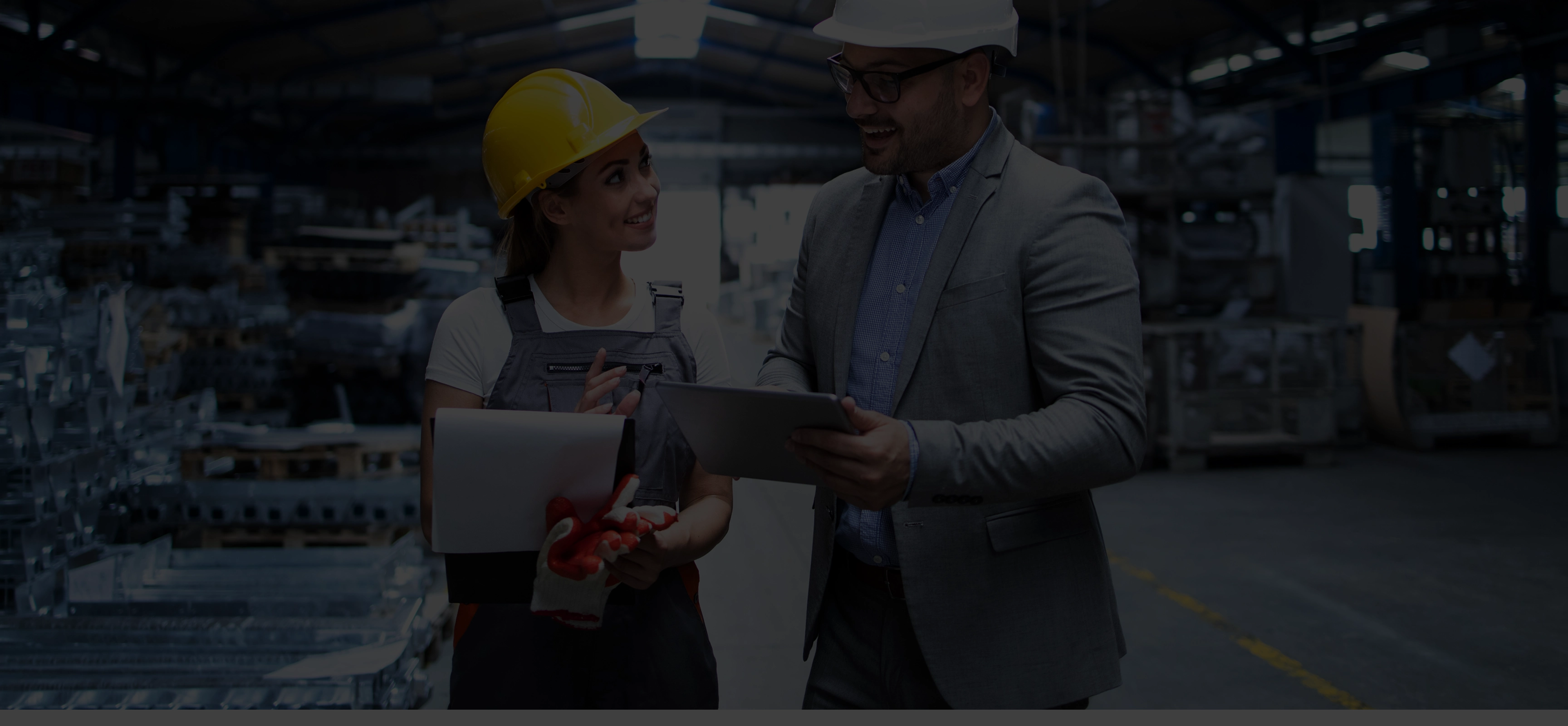 Woman in yellow hard hat and work overalls holding papers and gloves talking to man in suit with white hard hat and tablet inside a manufacturing warehouse.
