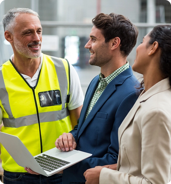 Three professionals in a discussion: a man in a high-visibility vest, a man in a blue suit holding a laptop, and a woman in a beige blazer.
