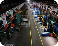 Industrial factory floor with rows of large blue machines and yellow overhead cranes along a central aisle marked with yellow lines.