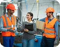Two male construction workers in orange safety vests and helmets talking with a woman holding a clipboard in an industrial setting.