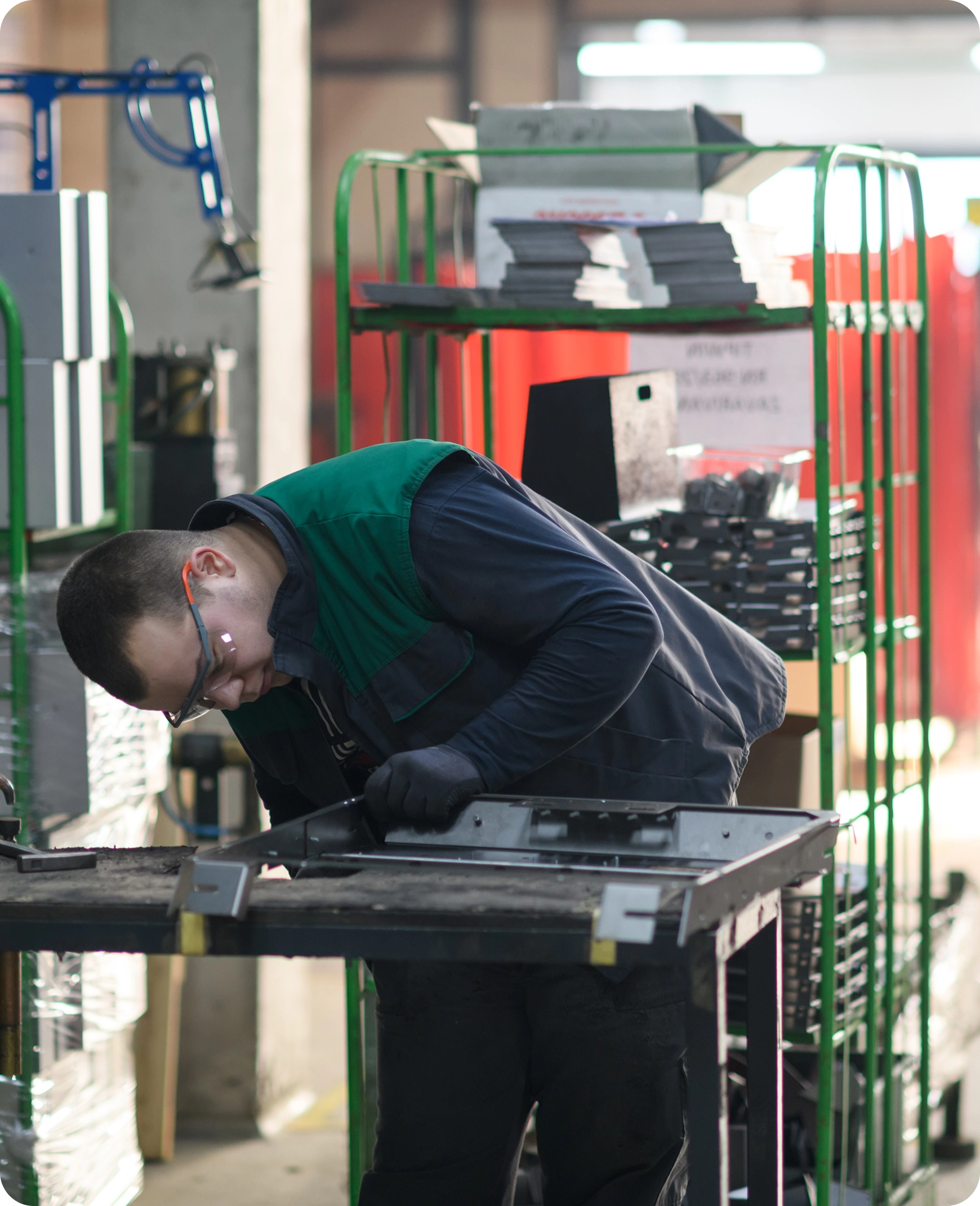 Worker wearing safety glasses and gloves inspecting a metal part on a workbench in an industrial setting.