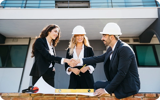Two businesswomen shaking hands with a businessman wearing hard hats at a construction site with a level tool in the foreground.
