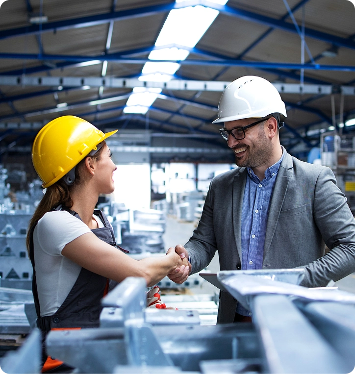 Male engineer in a suit and white hard hat shaking hands with female worker in a yellow hard hat inside a factory warehouse.