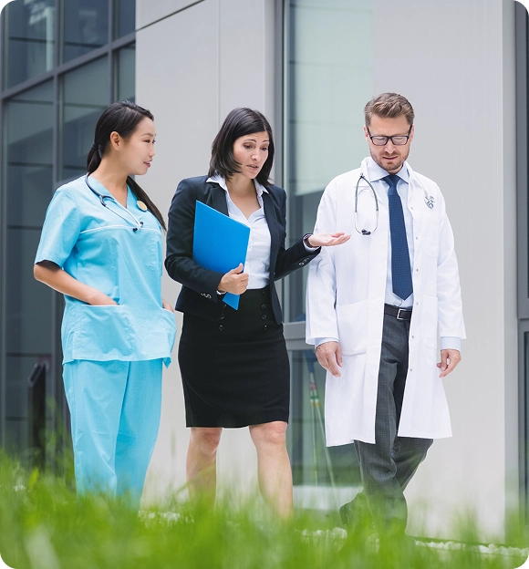 A nurse in blue scrubs, a businesswoman holding a blue folder, and a male doctor in a white coat walking outdoors near a modern building.