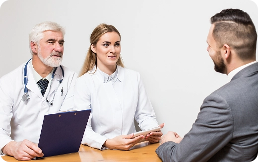 Two doctors in white coats consulting with a male patient in a gray suit across a wooden table.