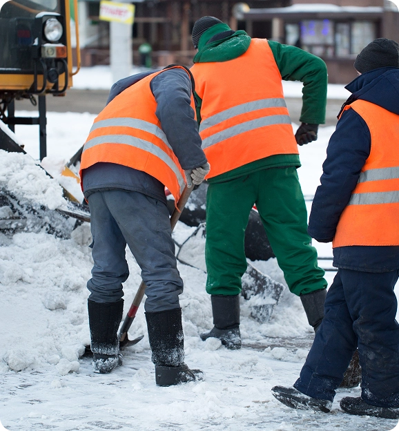 Three workers in orange reflective vests shoveling snow on a snowy street.